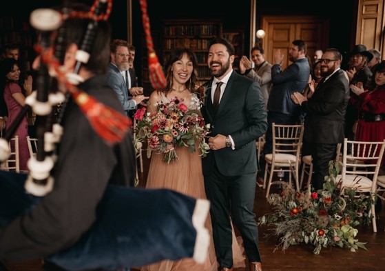 At a wedding in Cambo House, bride carries a natural bouquet of autumnal flowers with lush greenery.  Floral arrangements on the floor enhance the elegant, garden-inspired atmosphere.